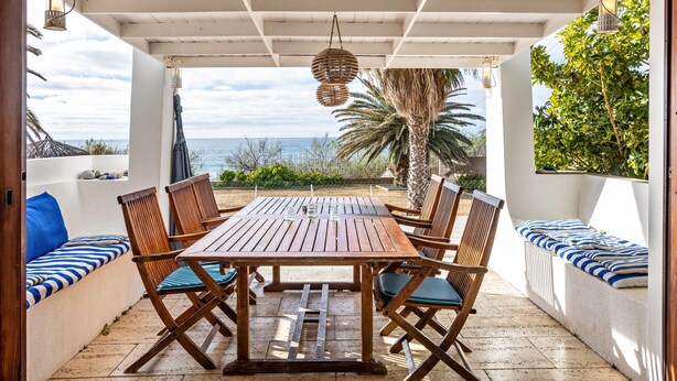 An outdoor patio with a wooden table and chairs, a view of the sea, and a white ceiling.