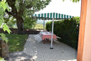 An outdoor patio with a checkered table and a striped umbrella, surrounded by greenery and a paved walkway. - Le Quiet Gîte à la Campagne (Charbonnières-les-Vieilles)