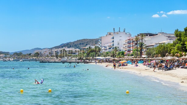 A beach with clear water, umbrellas, and buildings in the background.