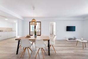 Une cuisine moderne avec des meubles blancs, une table à manger avec des chaises, un luminaire suspendu et un téléviseur à écran plat fixé au mur.