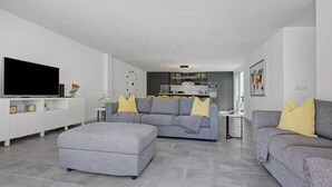 A modern living room with grey sofas, a grey ottoman, a flat-screen TV, and a kitchen in the background. - 'Villa Pacifica' with heated pool, terrace, lounge area & Wi-Fi (Corralejo)