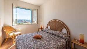 Ein Schlafzimmer mit Bett, einem Korbsessel, einem Nachttisch mit Kerze und Blick aufs Meer durch ein Fenster.