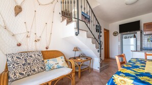 Ein Wohnzimmer mit einer Couch, einem Esstisch mit einer Blumen Tischdecke, einem kleinen runden Tisch mit einer Lampe, einer Treppe und einer Küchenzeile inklusive Kühlschrank.