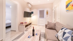 Ein modernes Wohnzimmer mit einer Couch, einem Couchtisch mit Flasche und Gläsern, einem an der Wand montierten Fernseher und einer Tür zu einem anderen Raum.