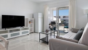 Ein modernes Wohnzimmer mit Flachbildfernseher, Glastisch, Sofa und einer Schiebeglastür, die zu einem Außenbereich führt.
