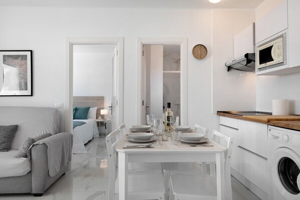 Une cuisine moderne équipée d’un lave-linge intégré, un coin repas avec une table pour quatre personnes et une chambre avec un lit et une table de chevet.