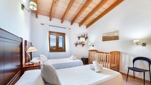 Un dormitorio con dos camas, una cuna, una silla, un ventanal con vistas, y un estante con libros y una lámpara.