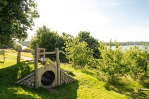 Ein Spielplatz mit einer hölzernen Kletterkonstruktion und einem Tunnel, umgeben von Gras und Bäumen, mit Blick auf ein Gewässer.