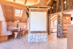 A wooden interior with a stone fireplace, a bench, a table with chairs, and a bed in the background.