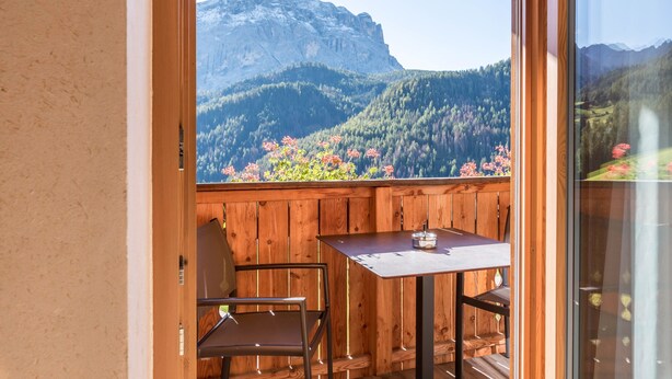 Ein Holzbalkon mit Tisch und Stühlen, mit Blick auf eine Berglandschaft mit dichten Wäldern und einem markanten Gipfel.