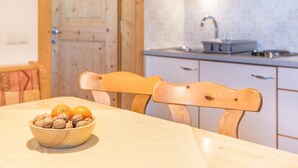 Ein Holztisch mit einer Obst- und Nuss-Schale, Holzstühle, ein Küchenbereich mit Spüle und Schränken sowie eine Holztür.