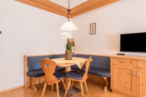 Un espace repas comprenant une table en bois, des bancs rembourrés de tissu bleu, un luminaire suspendu et un téléviseur posé sur un meuble en bois.