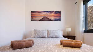 Un lit bien fait, avec une literie à motifs, un tableau représentant un coucher de soleil sur une jetée, et une fenêtre avec des rideaux.