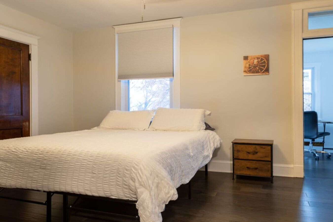 Un dormitorio con una cama, una mesita de noche de madera, una ventana con persianas y una puerta.