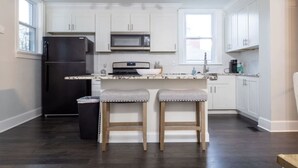Une cuisine moderne avec des meubles blancs, un réfrigérateur noir, un four à micro-ondes et une machine à café. On trouve deux tabourets en bois au comptoir.