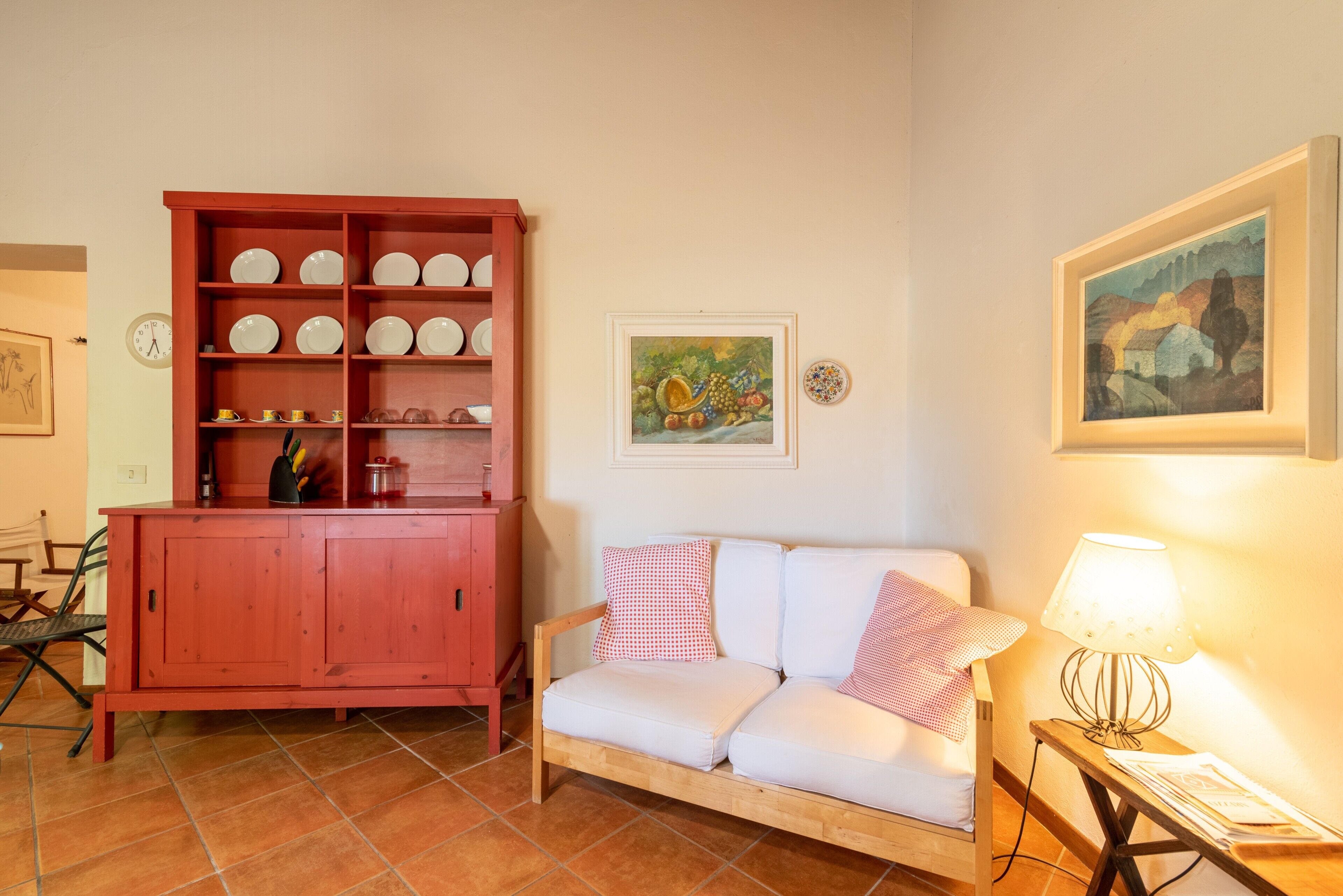 A room with a wooden sofa, a red cabinet with dishes, a framed painting, and a table lamp.