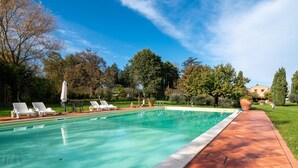 Une piscine avec des chaises longues, une terrasse carrelée et des arbres tout autour.