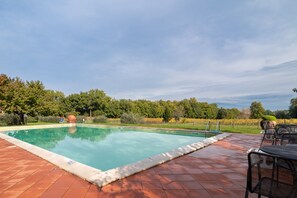 Una piscina rectangular con solárium de azulejos, rodeada de árboles y un cielo despejado.