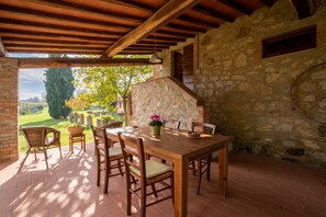 Une terrasse couverte avec une table et des chaises en bois, des murs en pierre et une vue sur un jardin.
