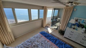 Une chambre avec une grande fenêtre donnant sur une plage, un lit recouvert d’une couvre-lit à motifs, une commode et un ventilateur de plafond.