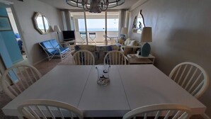 Un coin repas avec une table et des chaises blanches, un téléviseur et une vue sur la mer grâce à de grandes fenêtres.