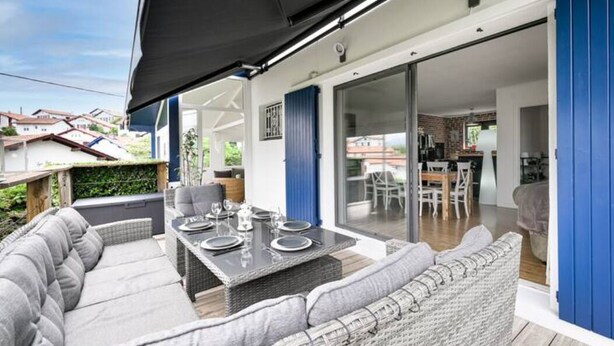 Un'area pranzo esterna coperta con un tavolo apparecchiato per quattro persone, adiacente a una porta scorrevole in vetro che dà su una cucina e sala da pranzo.