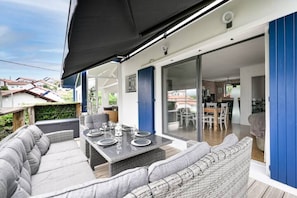 A covered outdoor dining area with a table set for four, adjacent to a sliding glass door leading to a kitchen and dining room. - Villa 8 Personnes Saint Jean de Luz à Pied (Ciboure)