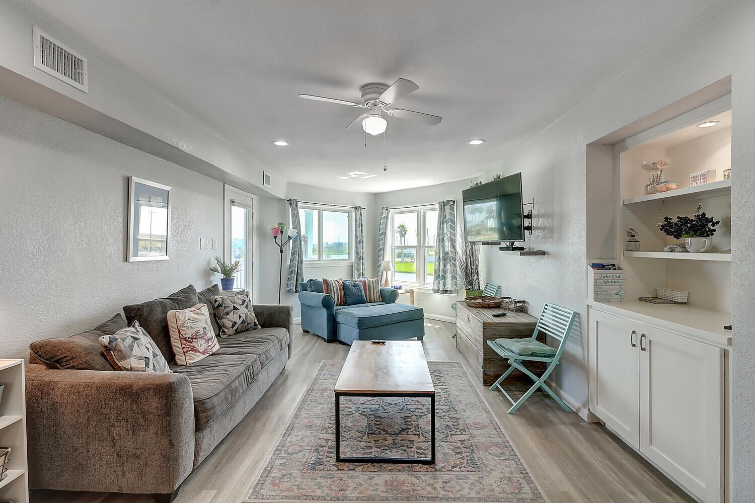 A spacious living room with a sofa, a coffee table, and a TV. There is a window with curtains and a ceiling fan.