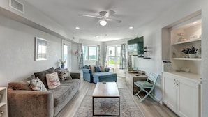 A spacious living room with a sofa, a coffee table, and a TV. There is a window with curtains and a ceiling fan.