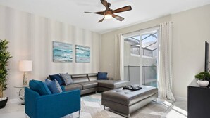 A modern living room with a sectional sofa, a coffee table, and a ceiling fan. There is a large window with curtains and a view of a building outside.