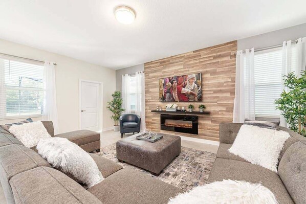 A modern living room with a fireplace, grey sofas, and a large wall-mounted TV. - Last Minute Disney! Private Pool, Hot Tub, Game Room & Themed Bedrooms. Book Now! #29466 (Orlando)