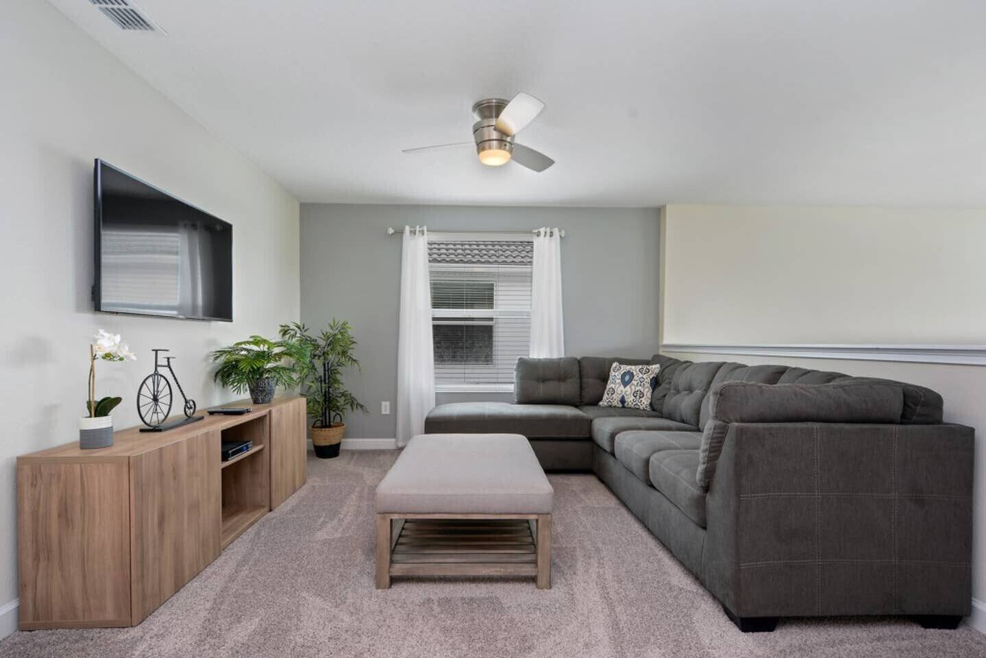 A modern living room with a grey sectional sofa, a wooden coffee table, a TV mounted on the wall, a ceiling fan, and a window with blinds.