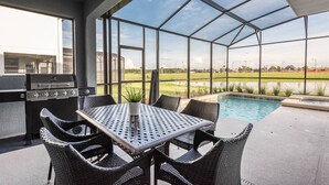 Un patio exterior con una parrilla, una mesa de comedor y sillas, con vista a una piscina y un área verde.