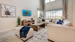 Amplia sala de estar con un sofá beige, una mesa de centro y un televisor en la pared. Se aprecia un ventanal con cortinas y vistas al exterior.