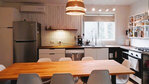 Moderni keittiö, jossa on puinen ruokapöytä, valkoiset tuolit, ruostumattomasta teräksestä valmistettu jääkaappi, astianpesukone, kaasuliesi ja ikkuna, josta tulee luonnonvaloa.