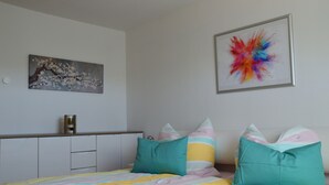 Un dormitorio con cama, almohadas, una mesita de noche y dos cuadros colgados en la pared.