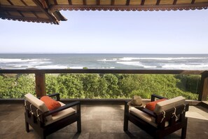 A balcony with two wooden chairs, one with a red cushion and the other with a straw hat. The balcony overlooks a view of the ocean and lush greenery.