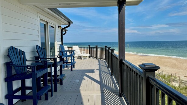 Una terraza de madera con sillas Adirondack azules, una mesa de madera y vista a la playa y el océano.