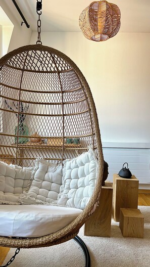 A hanging chair with a woven frame and white cushions, wooden blocks, and a woven light fixture in a room with a carpeted floor. - Le Verdoyant *** (Colmar)