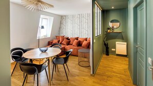 A modern living room with a wooden floor, a round dining table with chairs, a sofa with cushions, and a ceiling light fixture. - Le Verdoyant *** (Colmar)