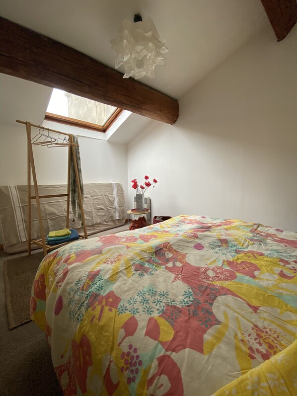 Un dormitorio con una colcha de patchwork colorida, un perchero de madera, un tragaluz y una mesita con un jarrón de flores.