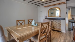 Uma sala de jantar com mesa e cadeiras de madeira, cozinha ao fundo e espelho sobre a pia.