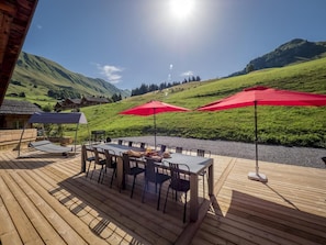 Een houten terras met een tafel en stoelen, twee rode parasols, en uitzicht op groene heuvels en een heldere lucht.