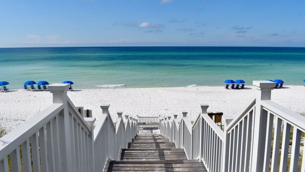 A wooden staircase leading to a beach with white railings and blue umbrellas.