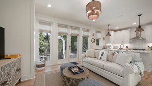 A spacious living room with a white sofa, patterned ottomans, a coffee table, and a large window with blinds.