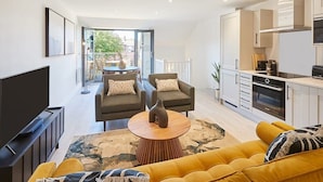 A modern living room with a sofa, armchairs, a coffee table, and a TV. There is a balcony with a table and chairs. The kitchen is visible with cabinets and appliances. - Host & Stay | St Johns Mews (York)