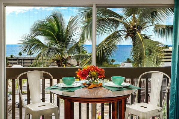 Un balcon avec une table dressée pour deux personnes, donnant sur une plage bordée de palmiers et sur l’océan.