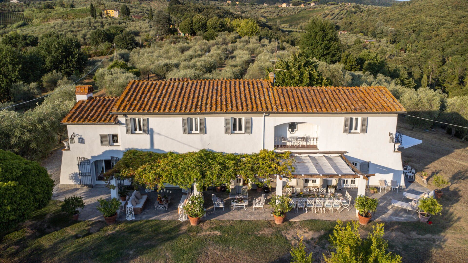 Una villa blanca con techo de tejas, un porche cubierto con mobiliario exterior y un jardín con plantas en macetas.