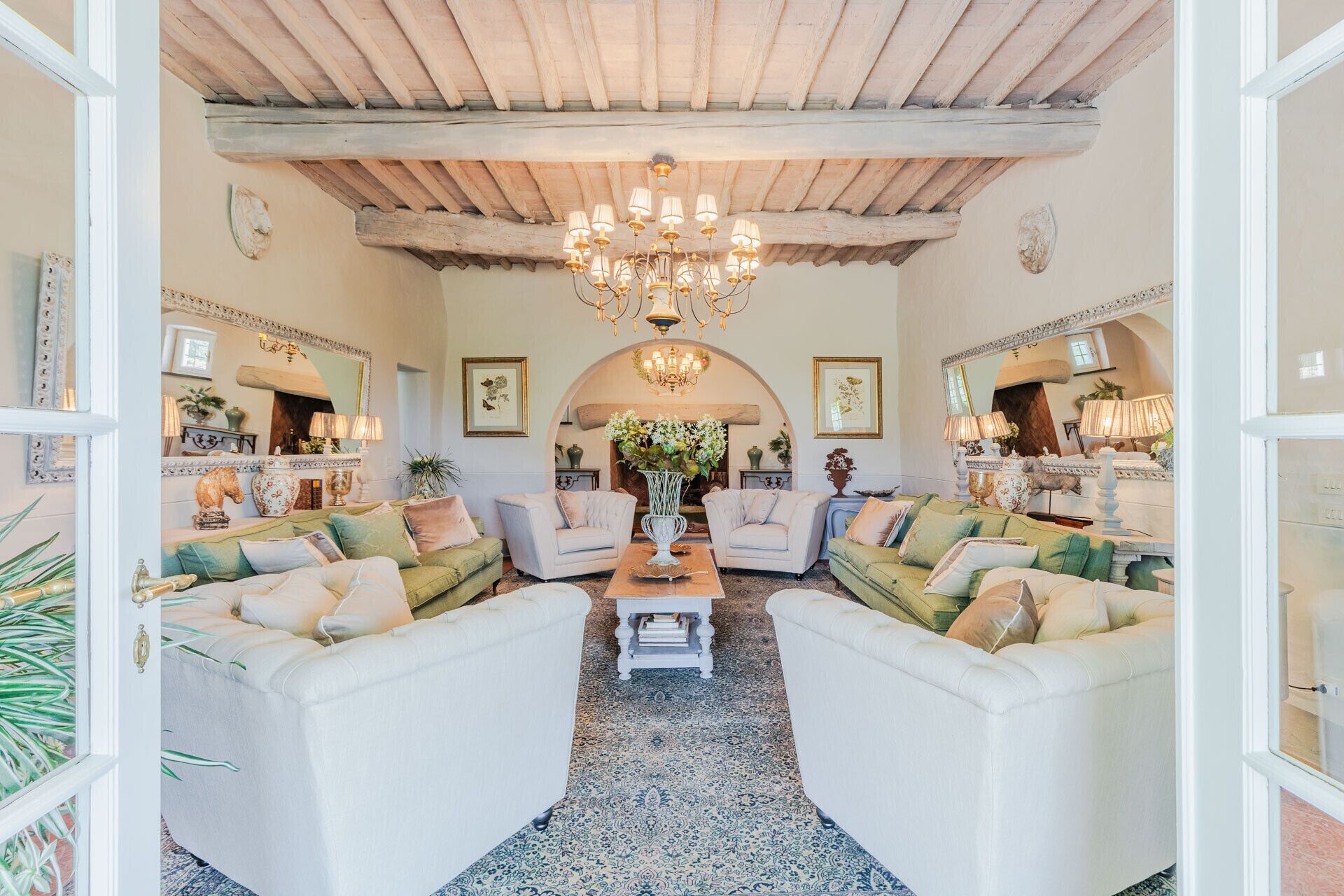 A spacious living room with a high ceiling, a chandelier, and a fireplace. There are two sofas and a coffee table in the center.