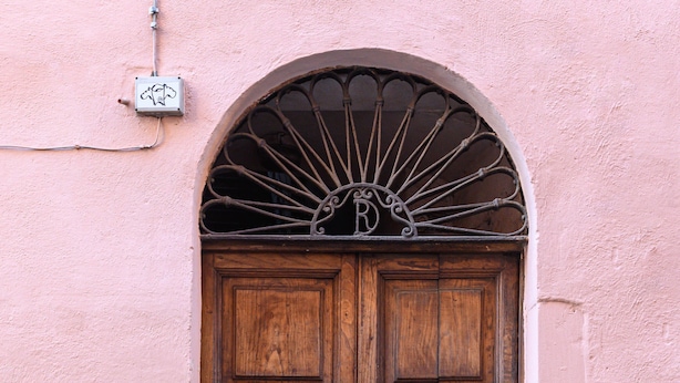 A wooden double door with a metal arch above it, set in a pink wall.